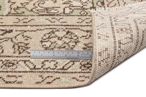 Load image 1 in gallery view. A close-up of the Mamail - Vintage Medallion Rug (6'4 x 9'7) shows its beige patterned surface with a folded corner revealing the back. A metal ruler highlights the rug’s edge, texture, and intricate medallion floral design.