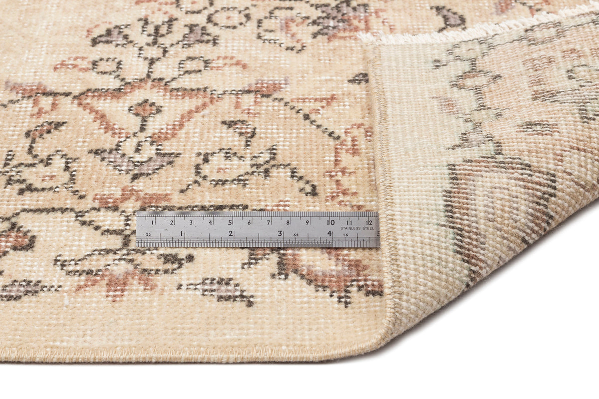 Beige rug with a faded floral medallion pattern and a metal ruler for scale, showing the underside and fringe.