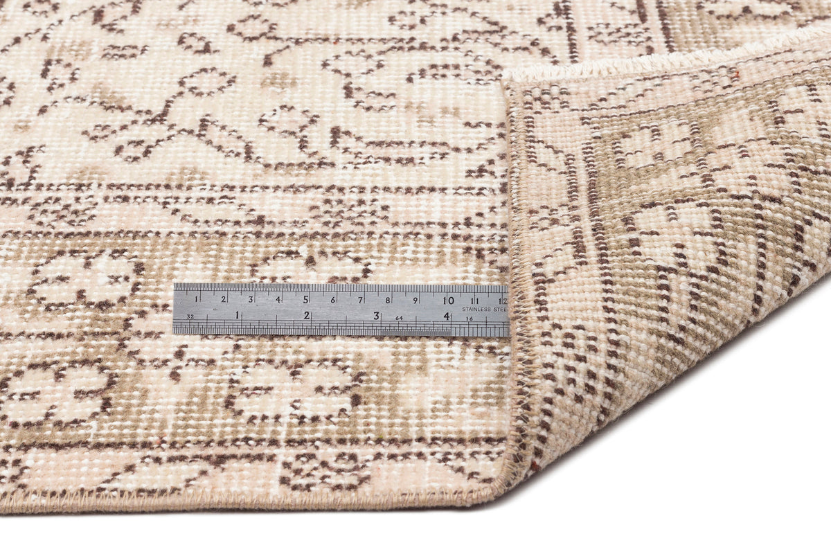 The Mofean - Vintage Floral Rug - 5'2 x 8'7 is shown in beige with brown floral flatweave designs. One corner is folded to reveal the underside, and a metal ruler (about 6 inches/15 cm) provides scale for this vintage hand-woven rug.