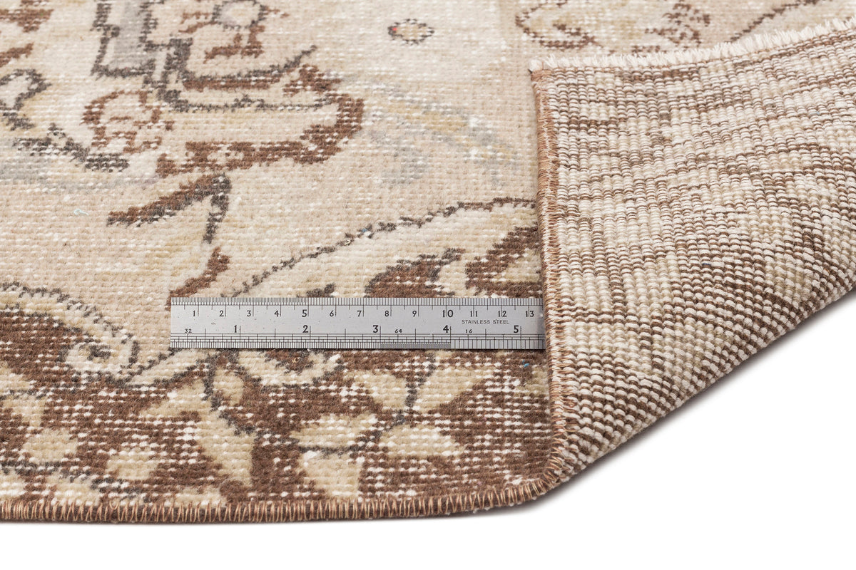 Beige and brown distressed floral rug with a folded corner revealing the woven backing and a metal ruler for scale.