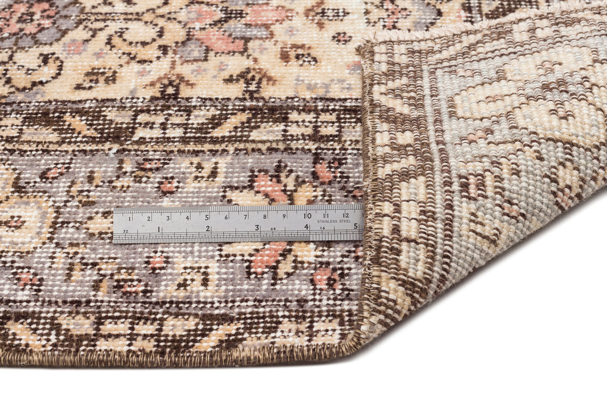 Beige, brown, and grey traditional floral rug with a folded corner showing the backing and a metal ruler for scale.