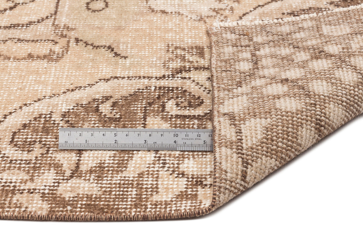Beige and brown distressed floral rug with a corner folded over to show the woven backing and a metal ruler for scale.