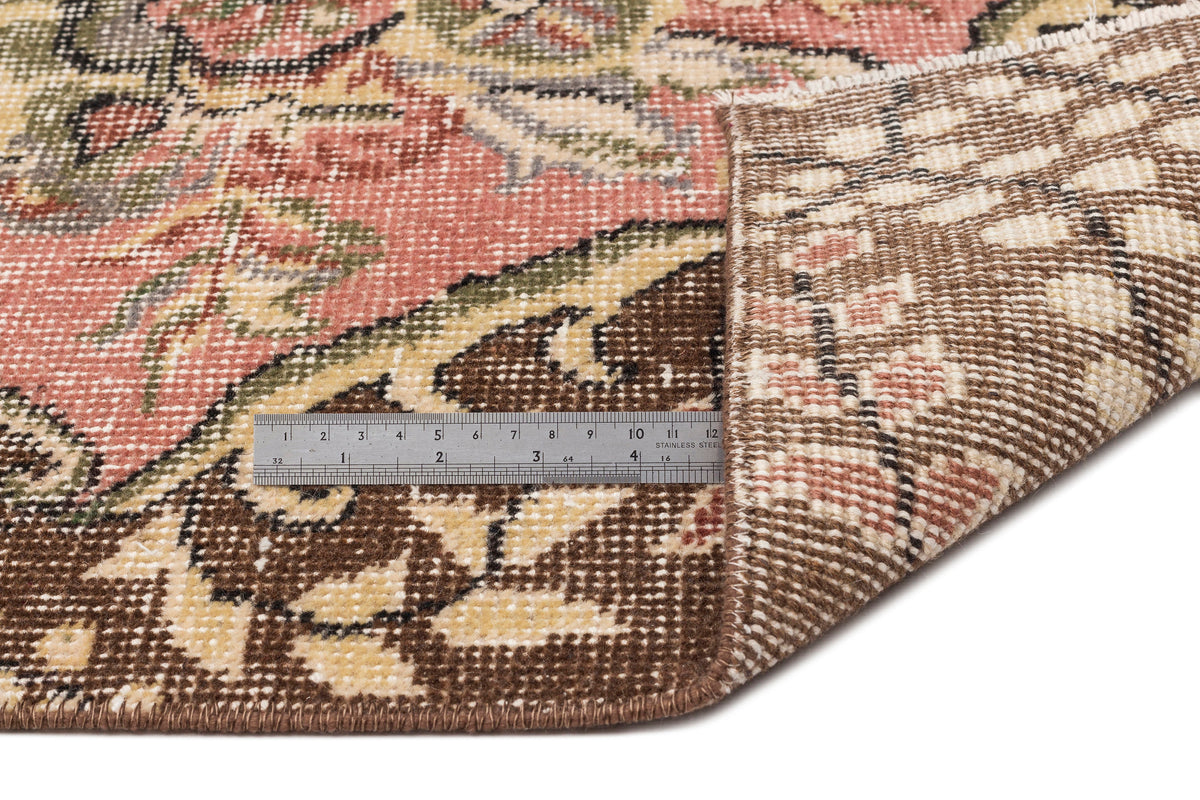 Pink, brown, and cream floral medallion rug with a folded corner revealing the woven backing and a metal ruler.