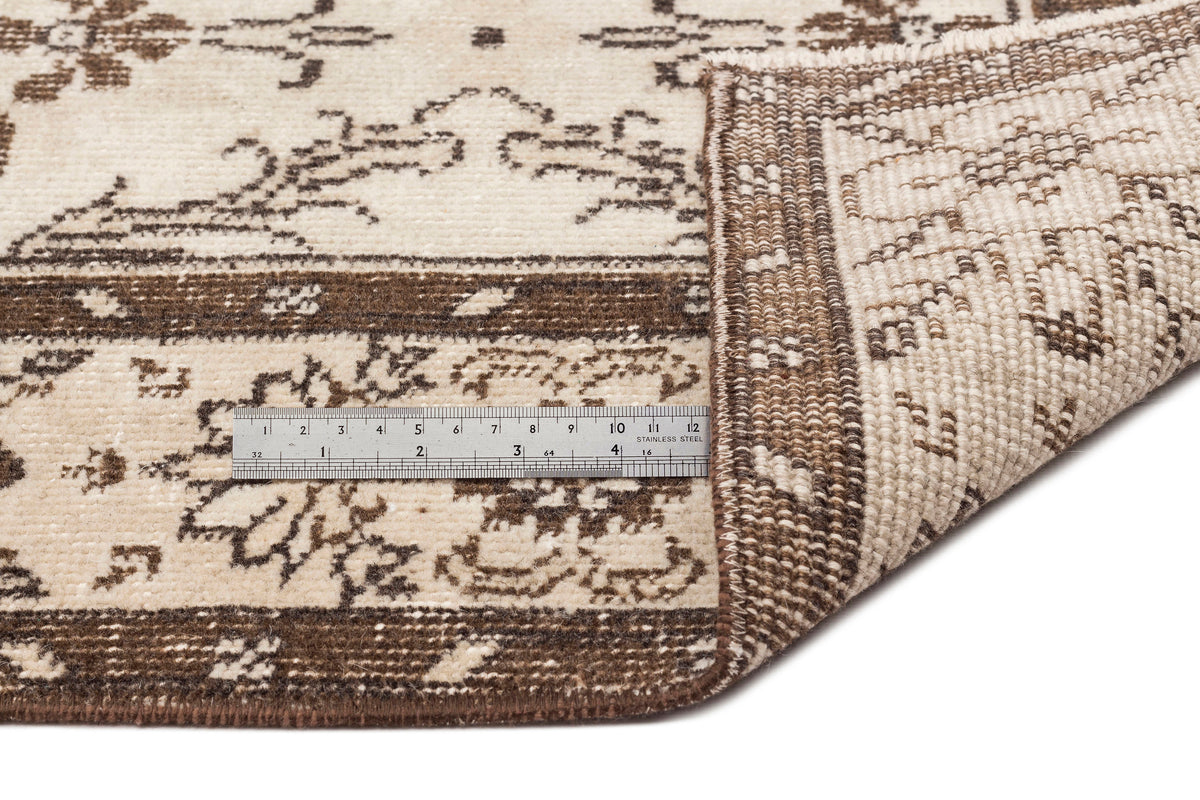Cream and brown rug with a traditional floral pattern, shown folded to reveal the backing and a metal measuring ruler.