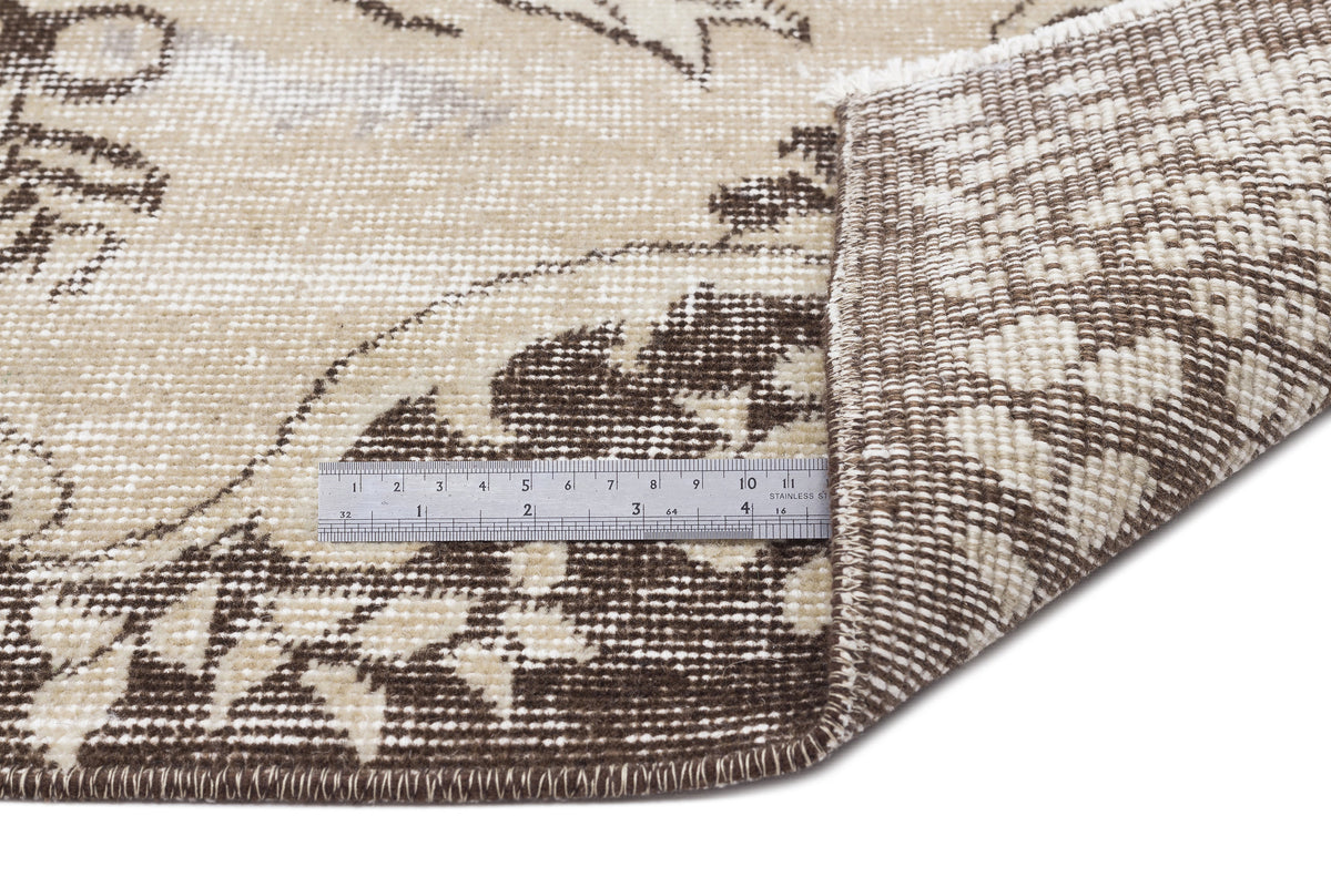 Beige and brown distressed floral rug with a folded corner showing the woven backing and a metal ruler for scale.