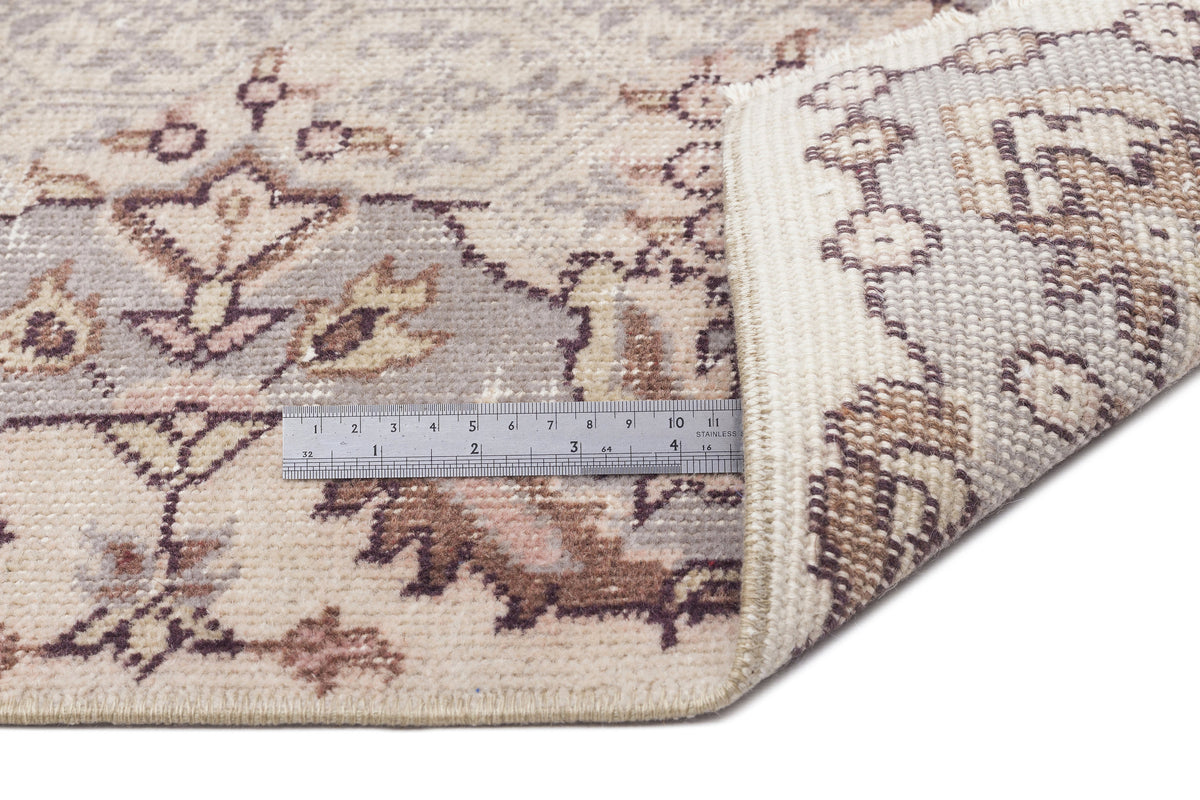 Beige and grey traditional floral rug with a corner folded back to show the weave, featuring a metal ruler for scale.