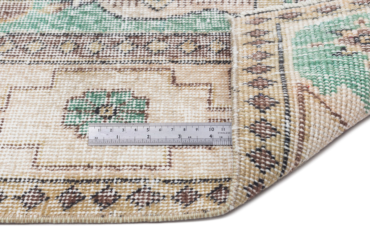Beige, green, and brown traditional patterned rug with a folded corner and a metal ruler for scale.