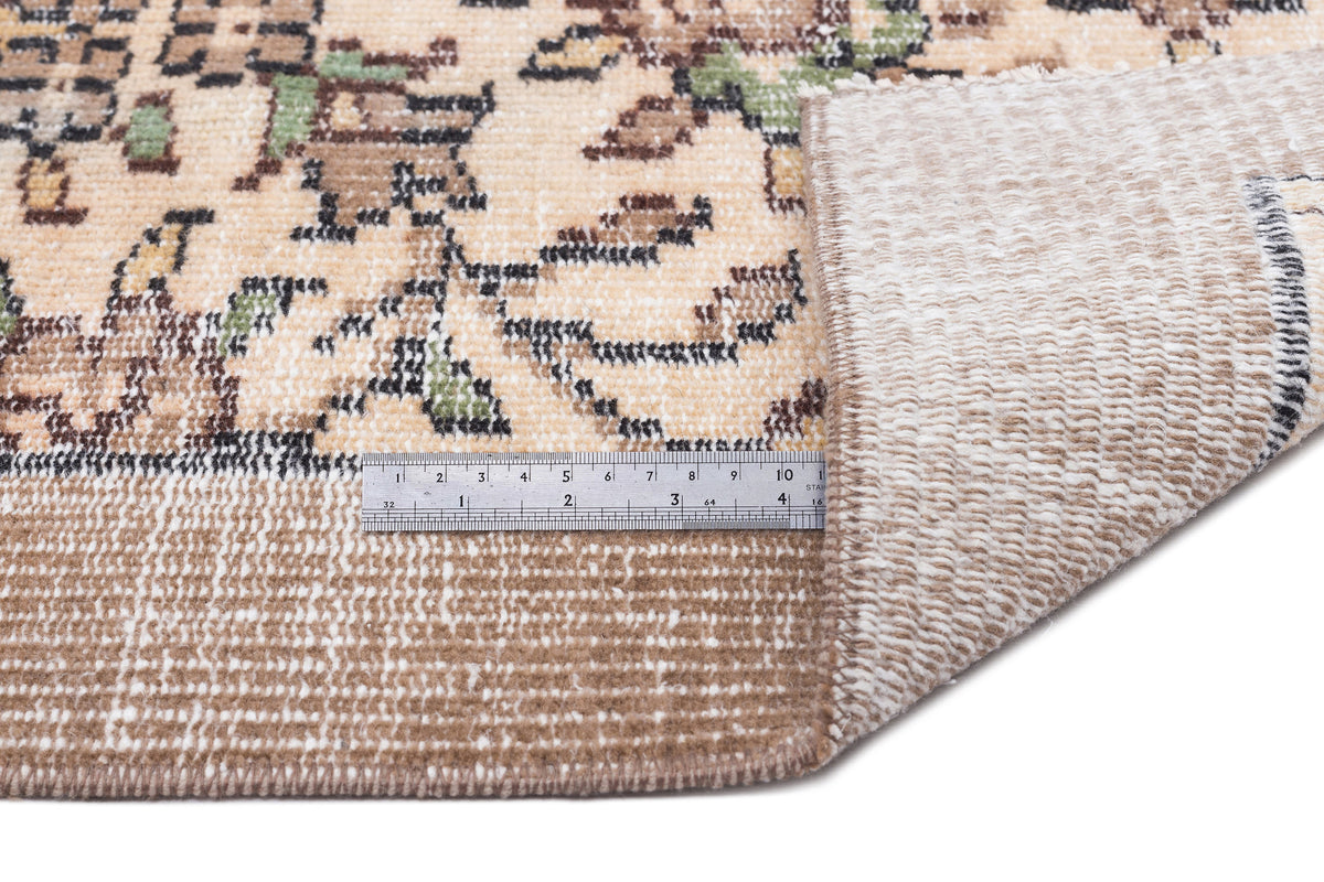 Beige rug with a distressed floral pattern and a metal ruler showing pile height, folded corner revealing the backing.