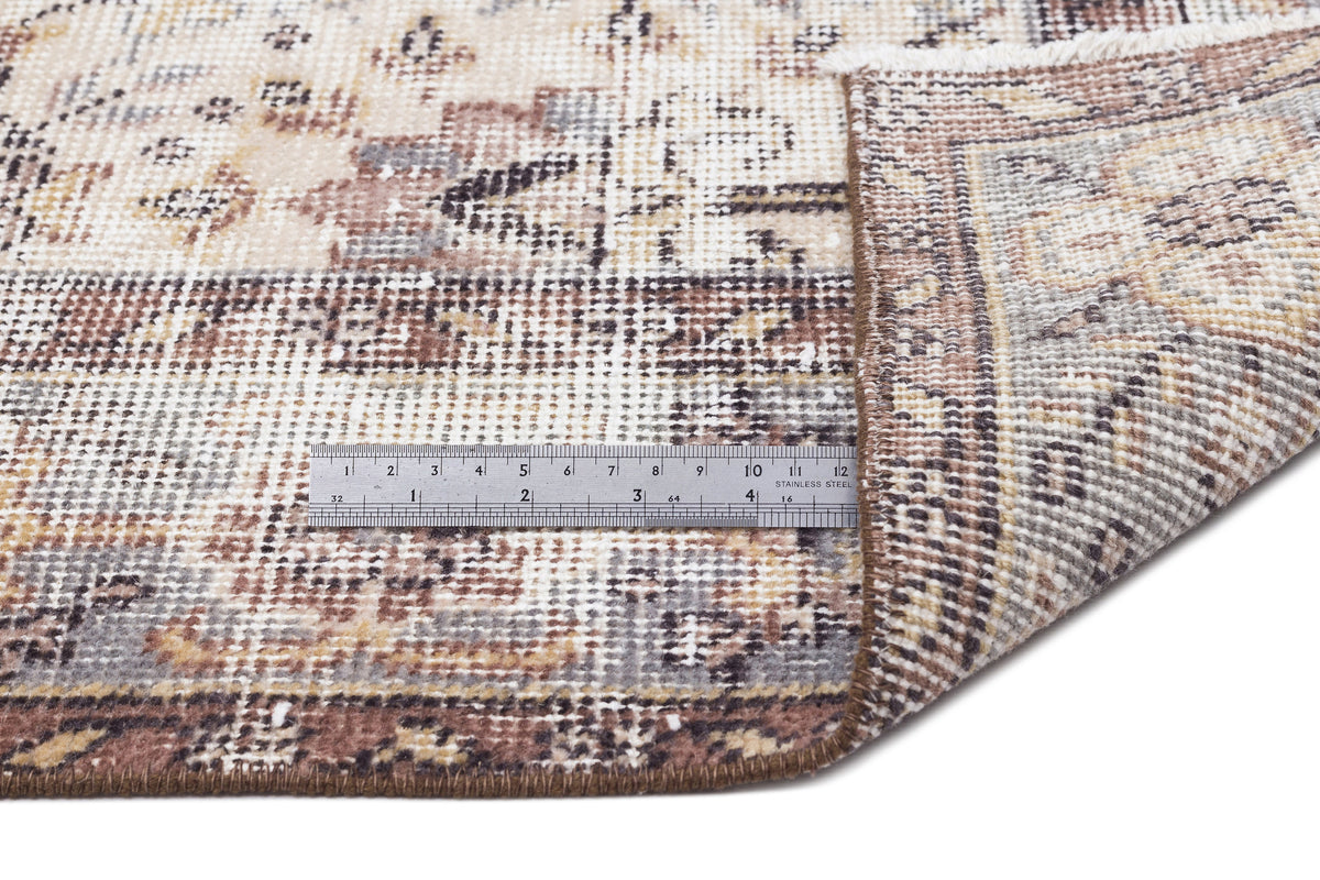 Distressed beige, brown, and grey traditional rug with a folded corner and a metal ruler for scale on a white background.