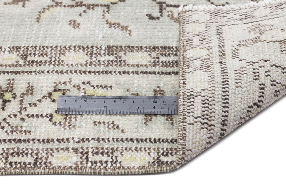 Sage green and cream rug with a distressed floral pattern, shown folded to reveal the underside with a metal ruler.
