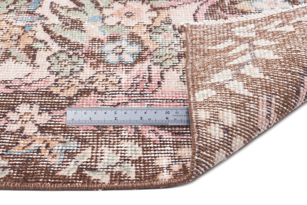 Distressed floral rug in brown, pink, and sage with a corner folded back to show the backing and a metal ruler for scale.