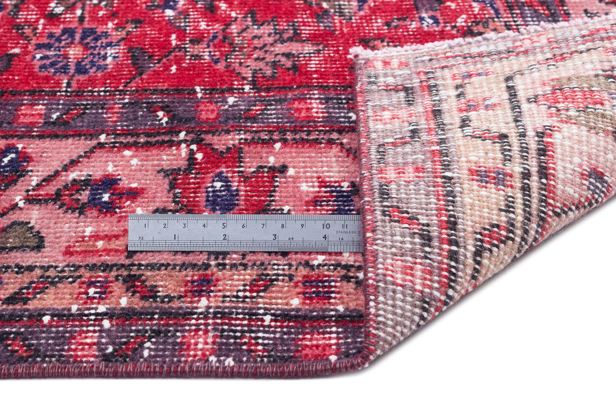 Distressed red and purple floral patterned rug with a folded corner revealing the beige backing and a metal ruler.