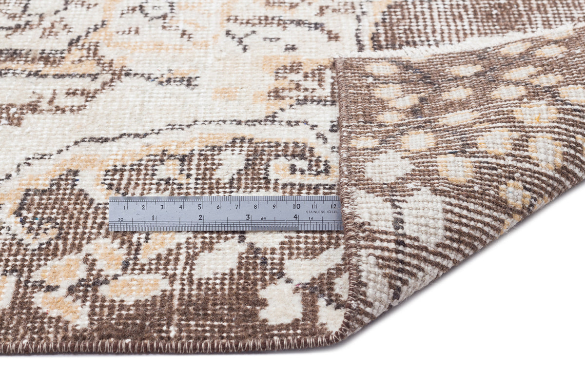 Brown and cream distressed medallion rug with a folded corner and a metal ruler for scale on a white background.
