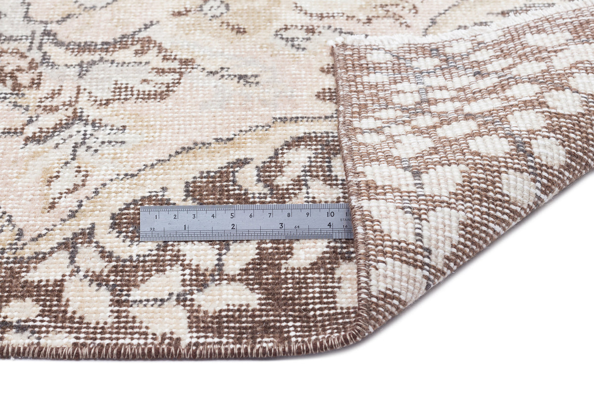Beige and brown distressed floral rug with a corner folded back, showing the woven backing and a metal ruler for scale.