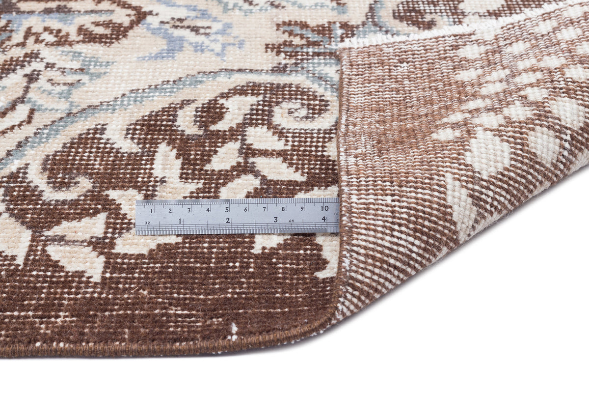 Brown, cream, and light blue floral medallion rug with a folded corner showing the woven backing and a metal ruler.