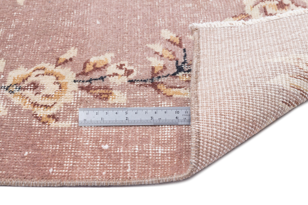 Dusty rose vintage rug with a cream and brown floral pattern, showing a corner fold and a metal ruler for scale.