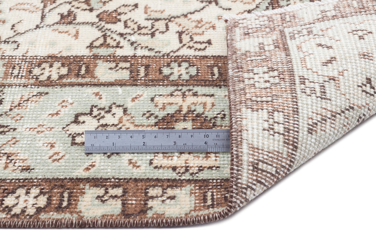 Beige, brown, and sage green traditional patterned rug with a folded corner and a metal ruler for scale.