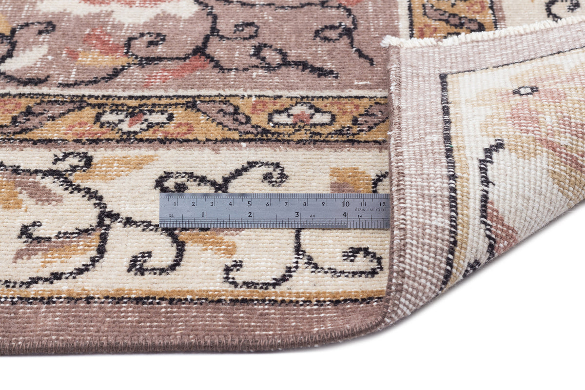 Brown and cream traditional rug with a floral border and a metal ruler showing pile height, isolated on white.