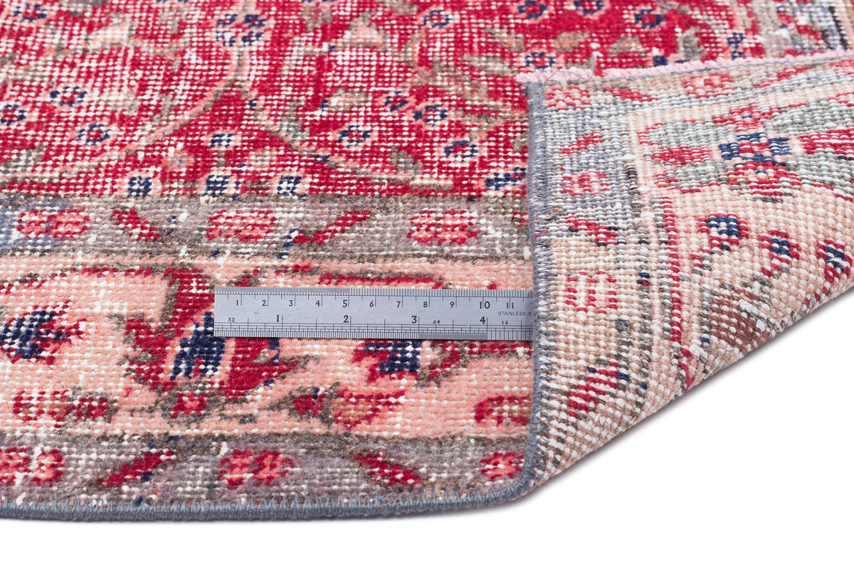 Red, pink, and blue distressed floral rug with a folded corner and a metal ruler for scale on a white background.