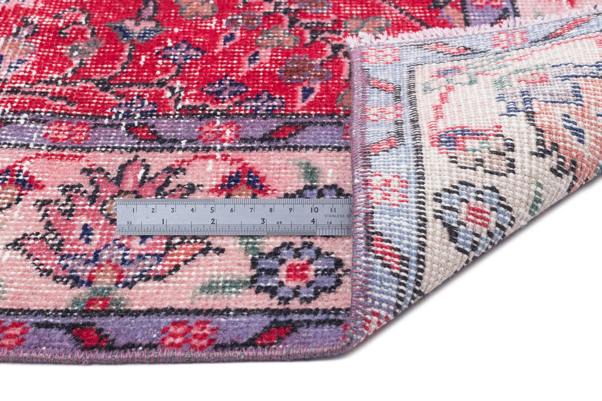 Red, pink, and purple floral patterned rug with a folded corner revealing a cream backing and a metal ruler for scale.