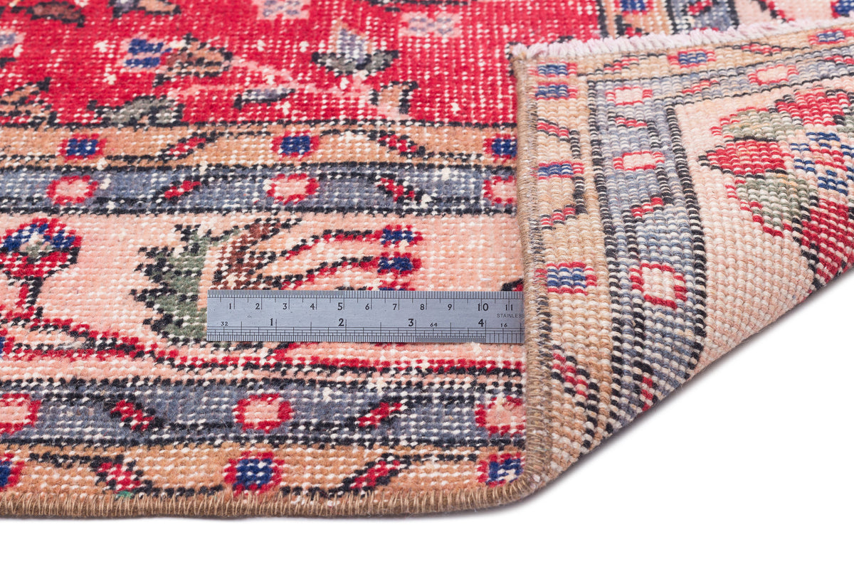 Red, peach, and blue traditional patterned rug with a folded corner and a metal ruler for scale on a white background.