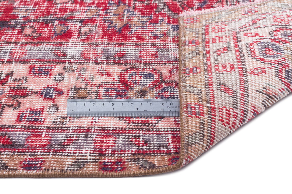 Red, pink, and grey distressed traditional rug with a folded corner and a metal ruler for scale on a white background.