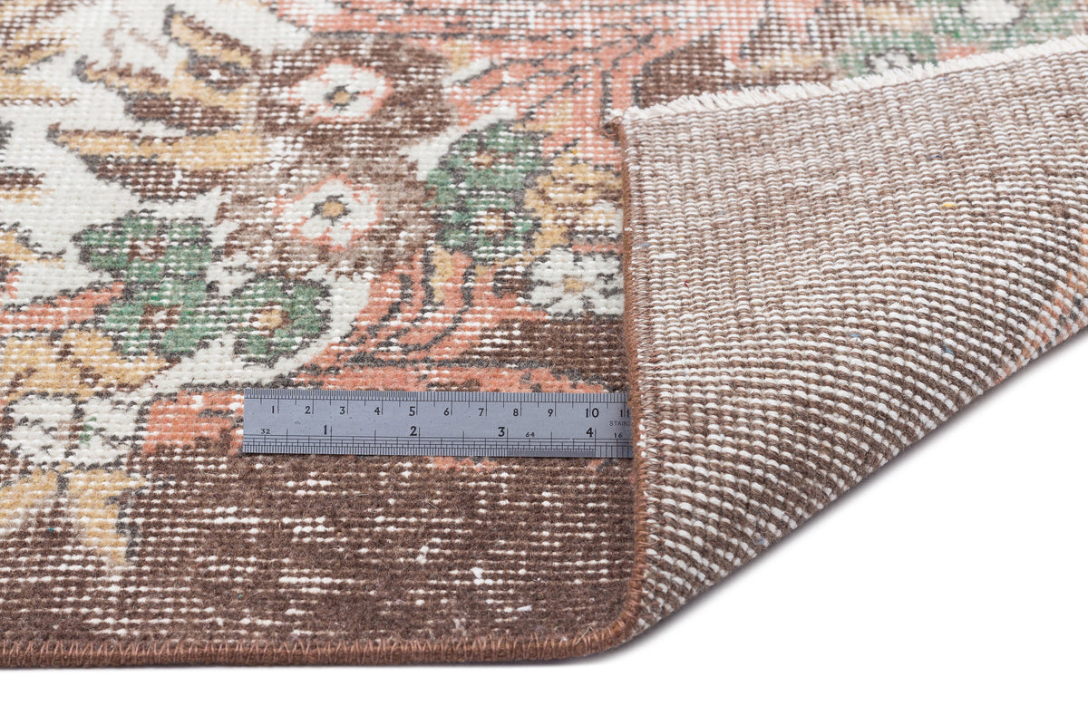 Distressed brown, cream, green, and orange floral medallion rug with a folded corner and a metal ruler for scale.
