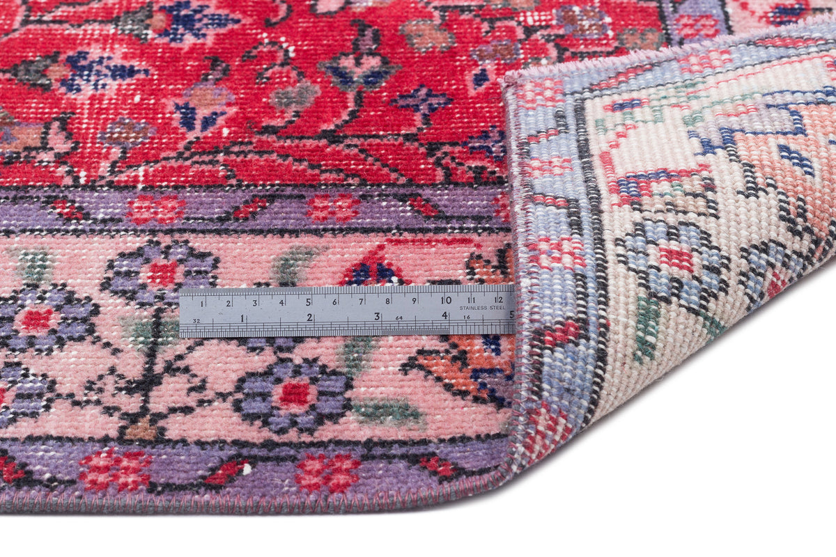 Red and pink traditional floral rug with purple accents, shown folded to reveal the woven backing and a metal ruler.