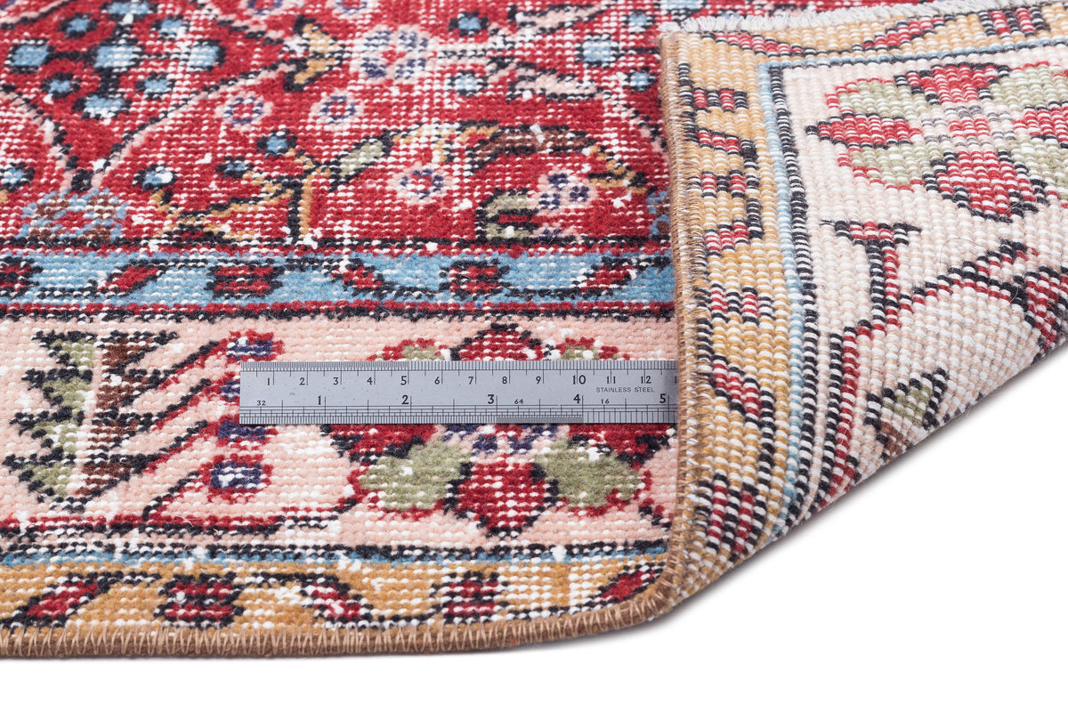 Red, pink, and blue traditional floral rug with a folded corner and a metal ruler for scale on a white background.