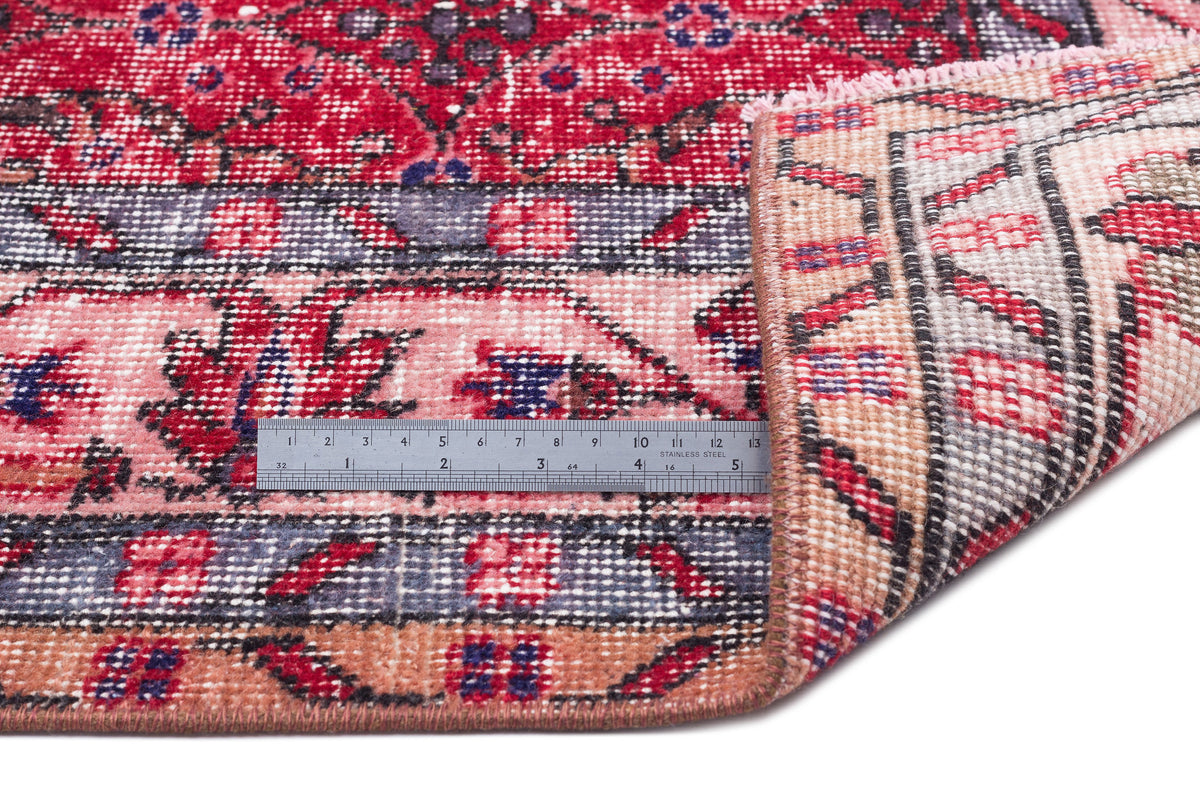 Red, pink, and blue distressed Persian-style rug with a floral pattern, shown with a metal ruler for scale.