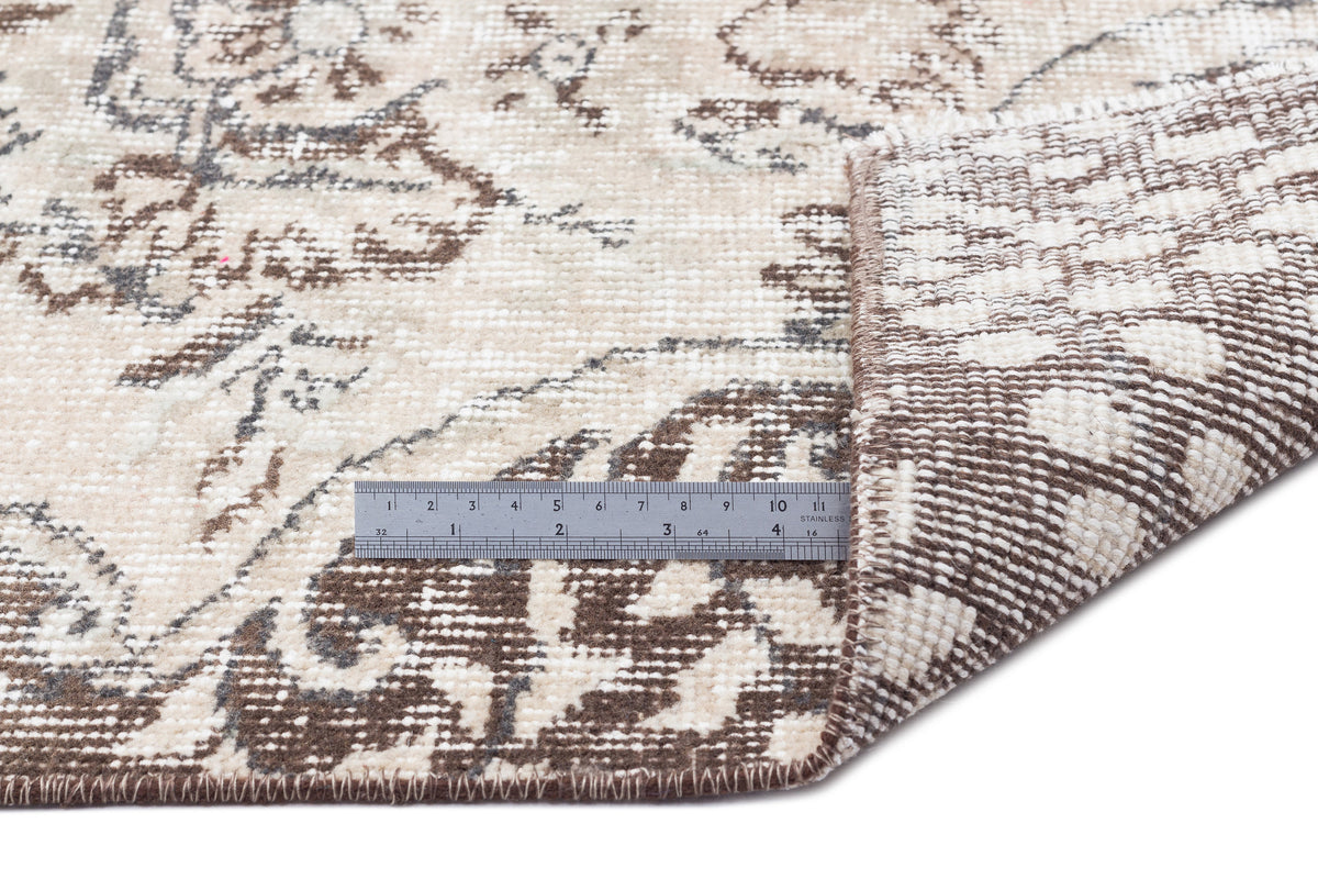 Beige and brown distressed floral rug with a folded corner and a metal ruler for scale on a white background.