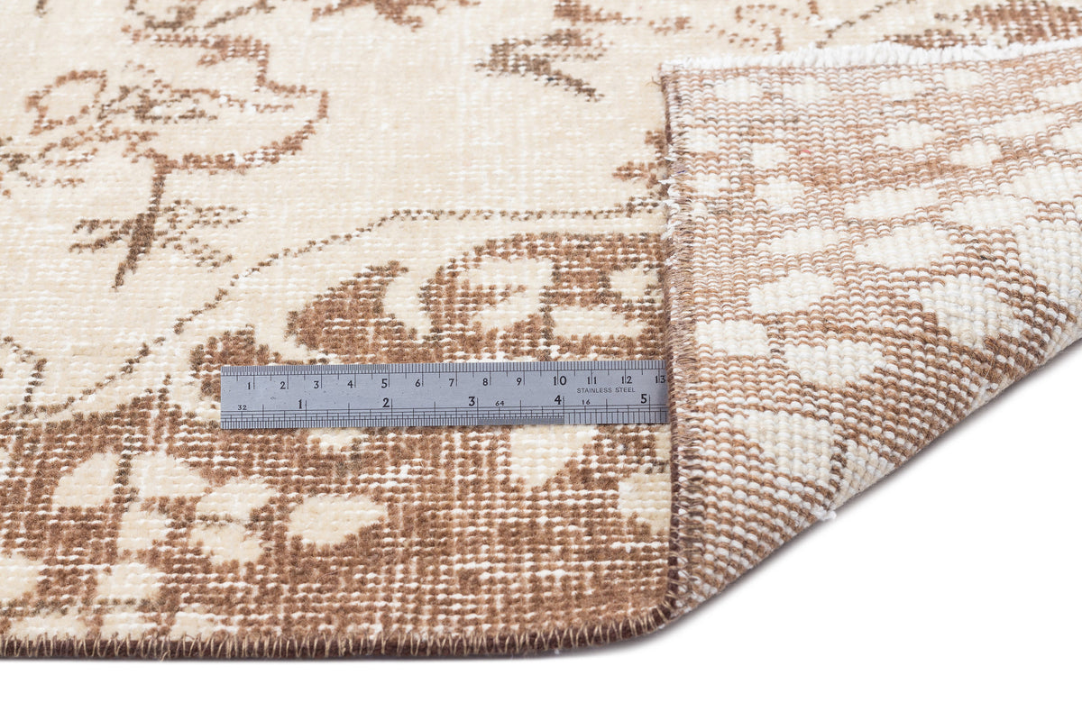 Beige and brown floral patterned rug with a folded corner showing the woven backing and a metal ruler for scale.
