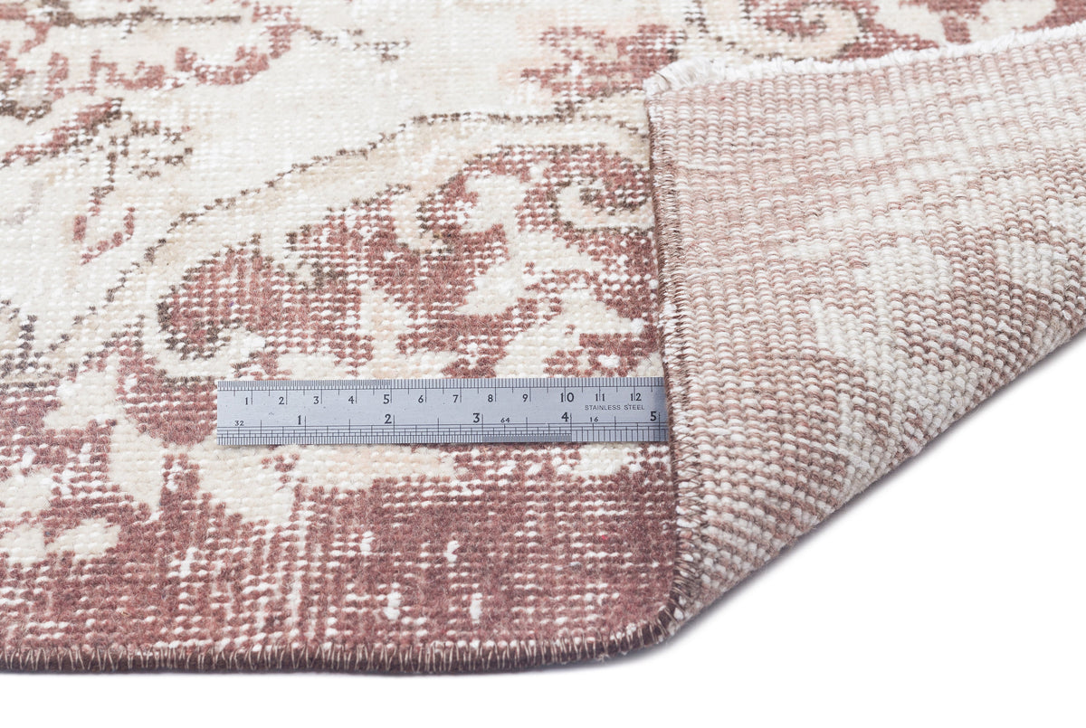 Cream and terracotta distressed floral rug with a folded corner and a metal ruler for scale on a white background.
