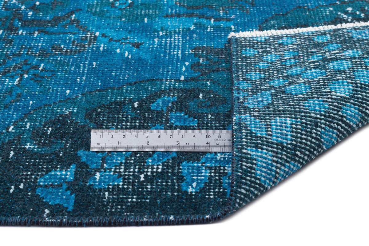 Distressed blue and black patterned rug with a folded corner revealing the woven backing and a metal ruler for scale.
