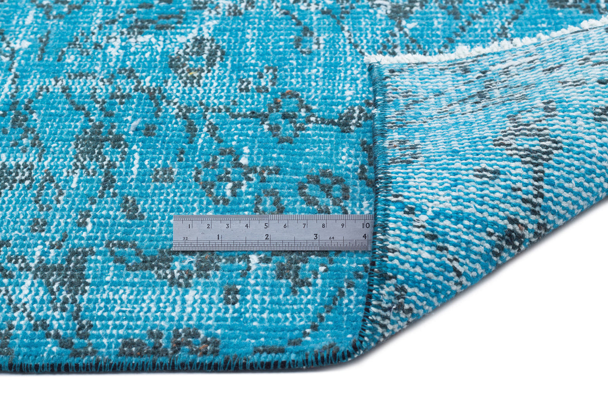 Bright turquoise rug with a distressed charcoal floral pattern, shown with a metal ruler for scale on a white background.