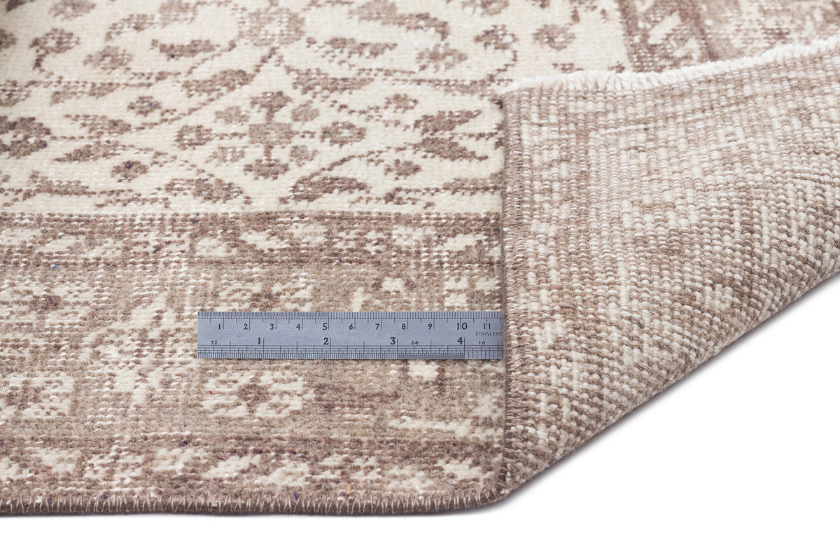 Beige and cream distressed floral rug with a folded corner and a metal ruler for scale on a white background.