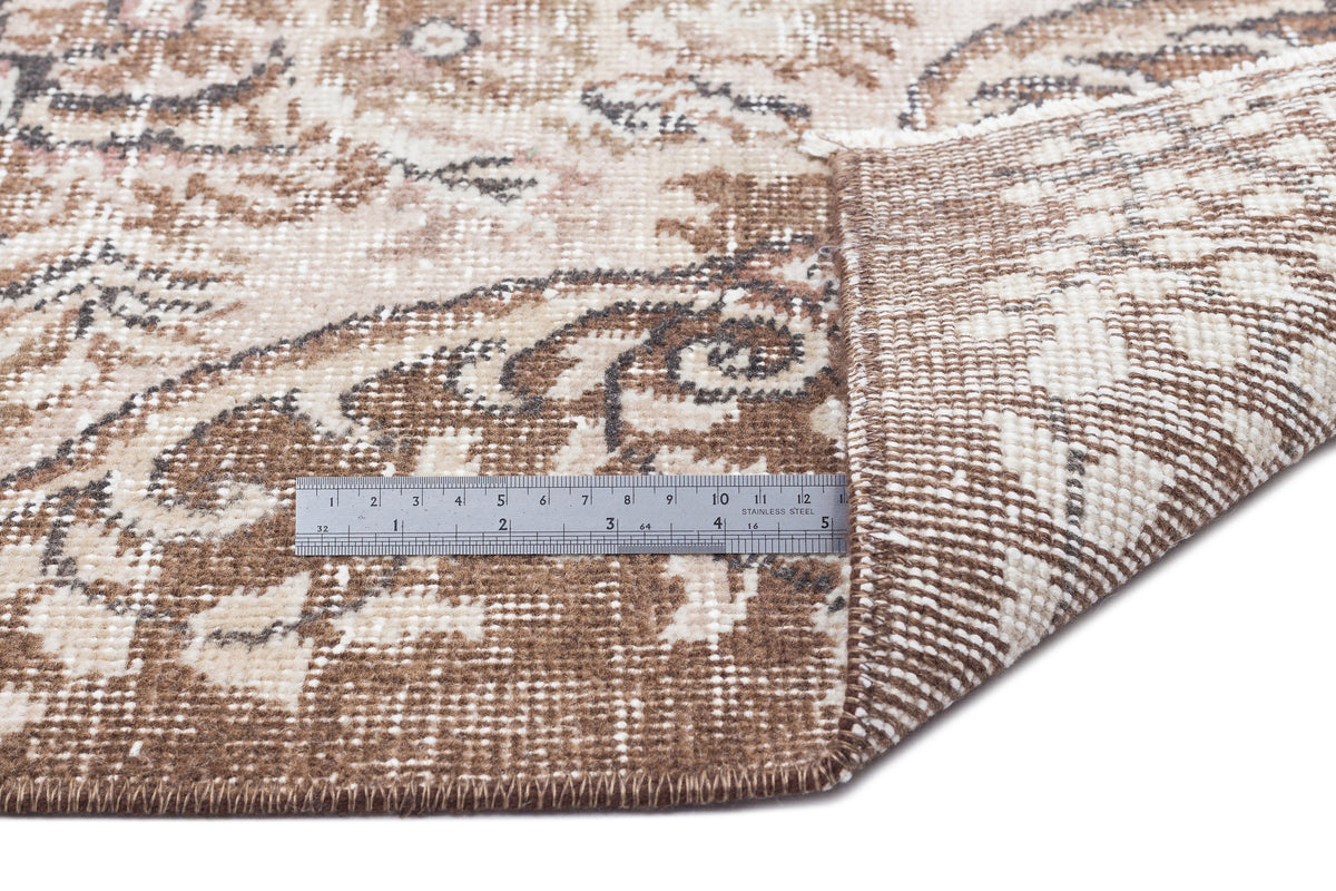 Brown and cream distressed floral medallion rug with a folded corner and a metal ruler for scale.