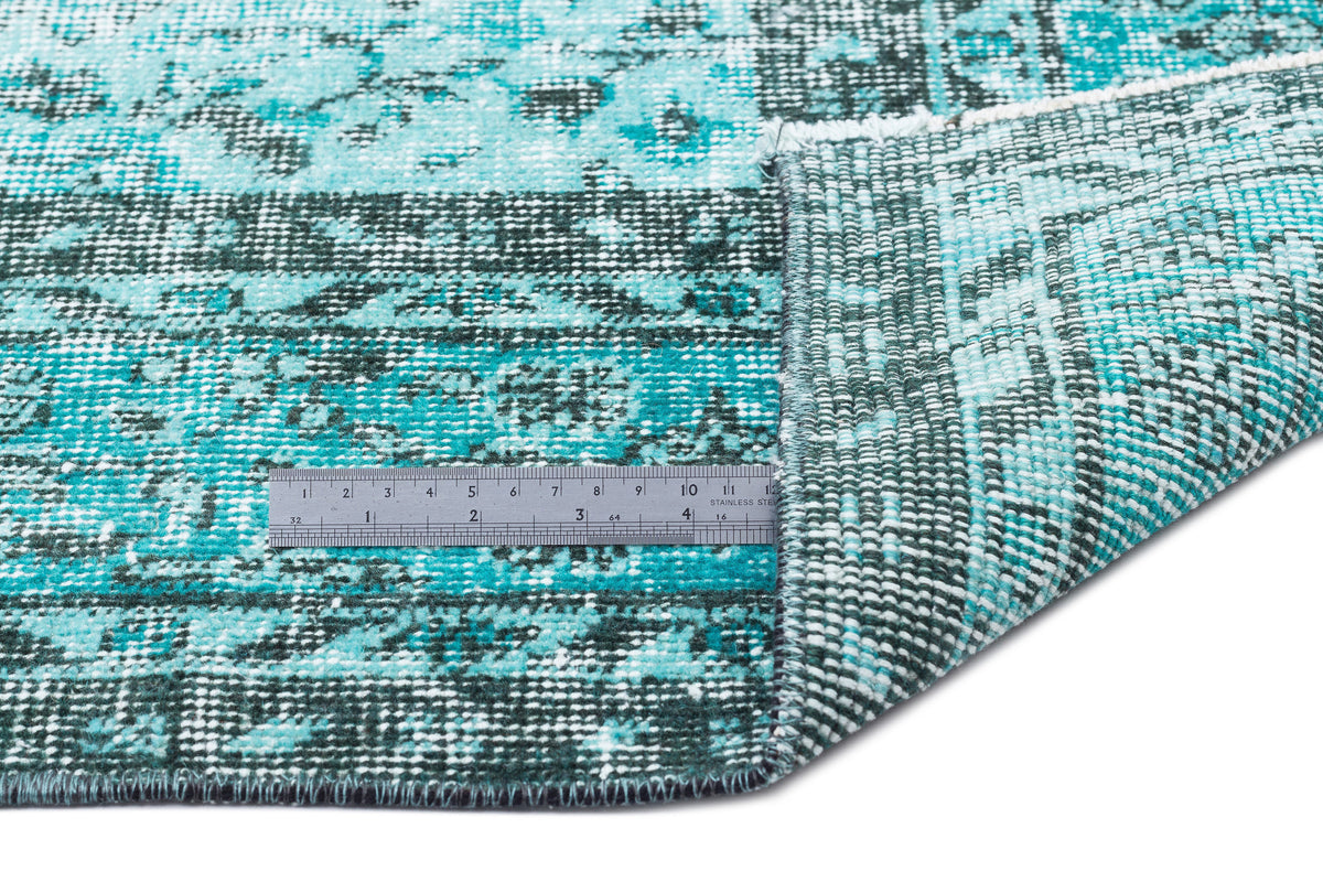 Turquoise and dark green distressed rug with a faded floral pattern, shown with a metal ruler for scale.