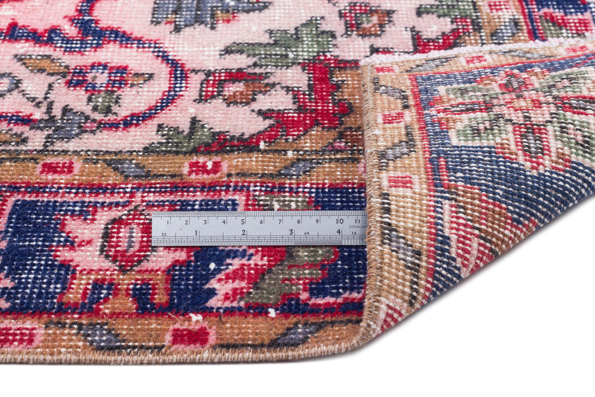 Pink, blue, and tan traditional Persian-style rug with a floral pattern, shown folded with a metal ruler for scale.