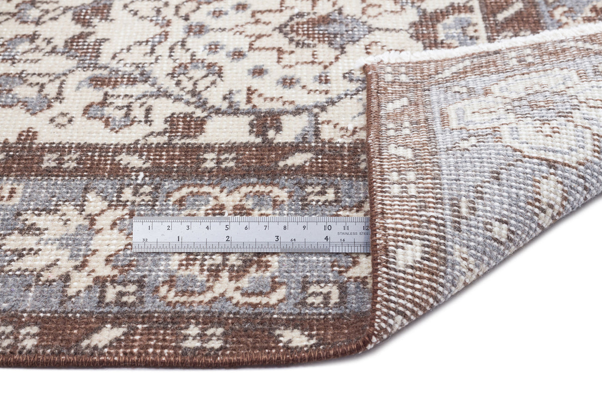 Brown, grey, and cream traditional floral rug with a folded corner and a metal ruler for scale on a white background.