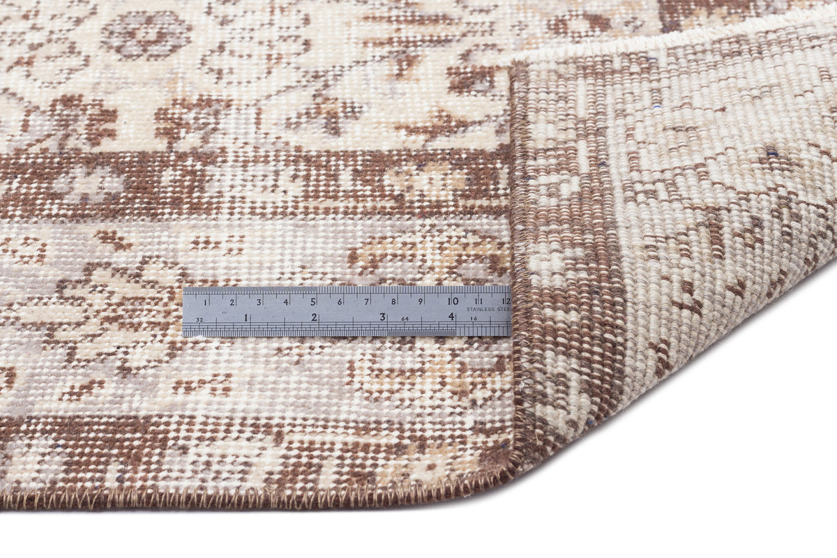Beige and brown distressed traditional rug with a floral pattern, shown with a metal ruler for scale on a white background.