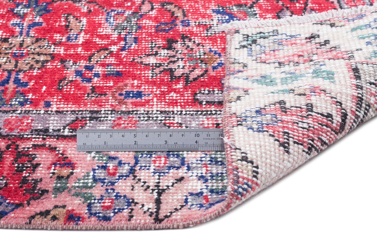 Red and pink traditional floral rug with a folded corner revealing the backing and a metal ruler for scale.