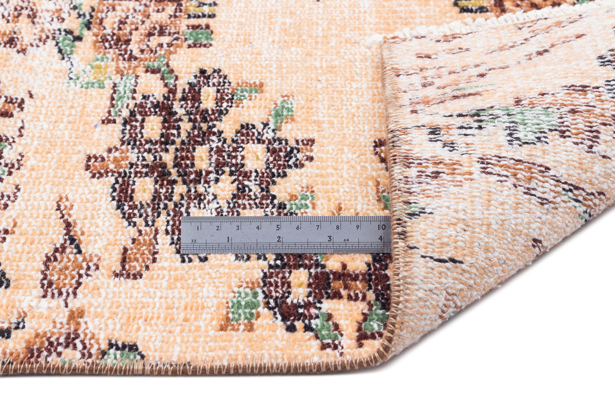 Peach, brown, and green floral patterned rug with a folded corner and a metal ruler for scale on a white background.