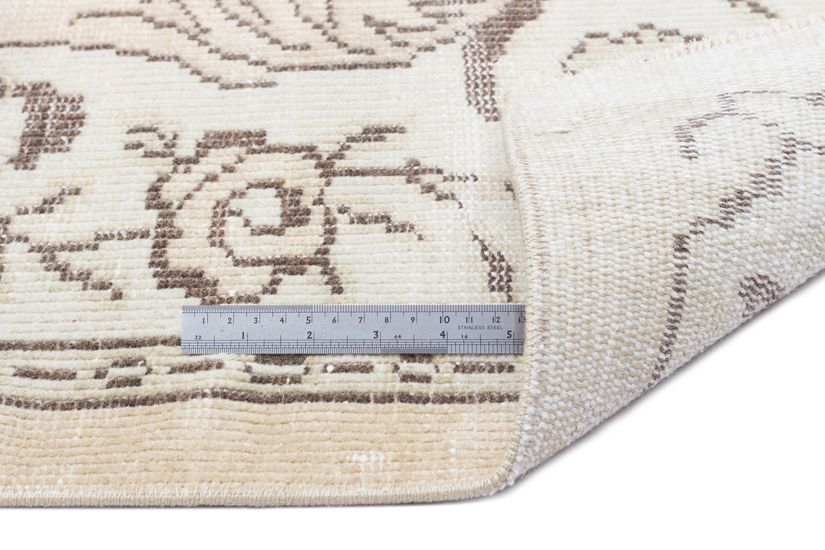 Cream and brown floral patterned rug with a folded corner and a metal ruler for scale on a white background.