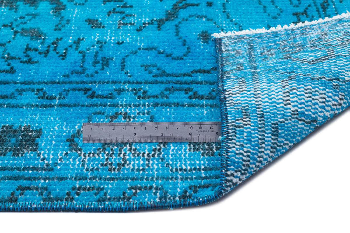 Bright blue rug with a distressed black floral pattern and a metal ruler showing scale, folded corner revealing backing.