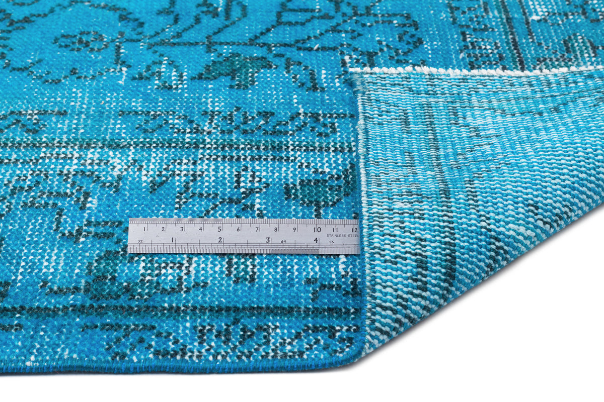 Bright blue distressed rug with a dark floral pattern, shown folded to reveal the woven backing and a metal ruler.