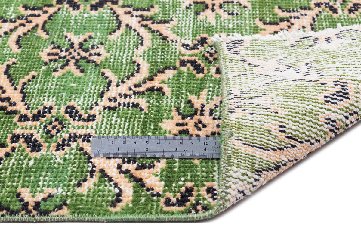 Green, black, and beige floral patterned rug with a folded corner and a metal ruler for scale on a white background.