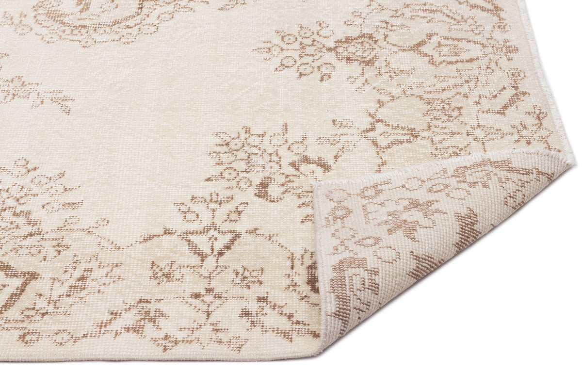 Cream and beige rug with a faded floral medallion pattern, shown with a corner folded over on a white background.