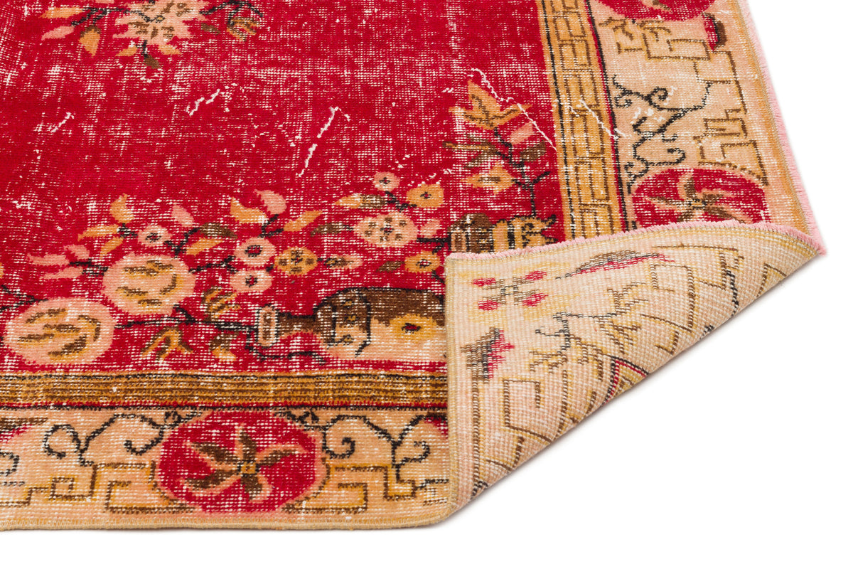 Distressed red rug with a floral pattern and beige geometric border, shown with a corner folded over on a white background.