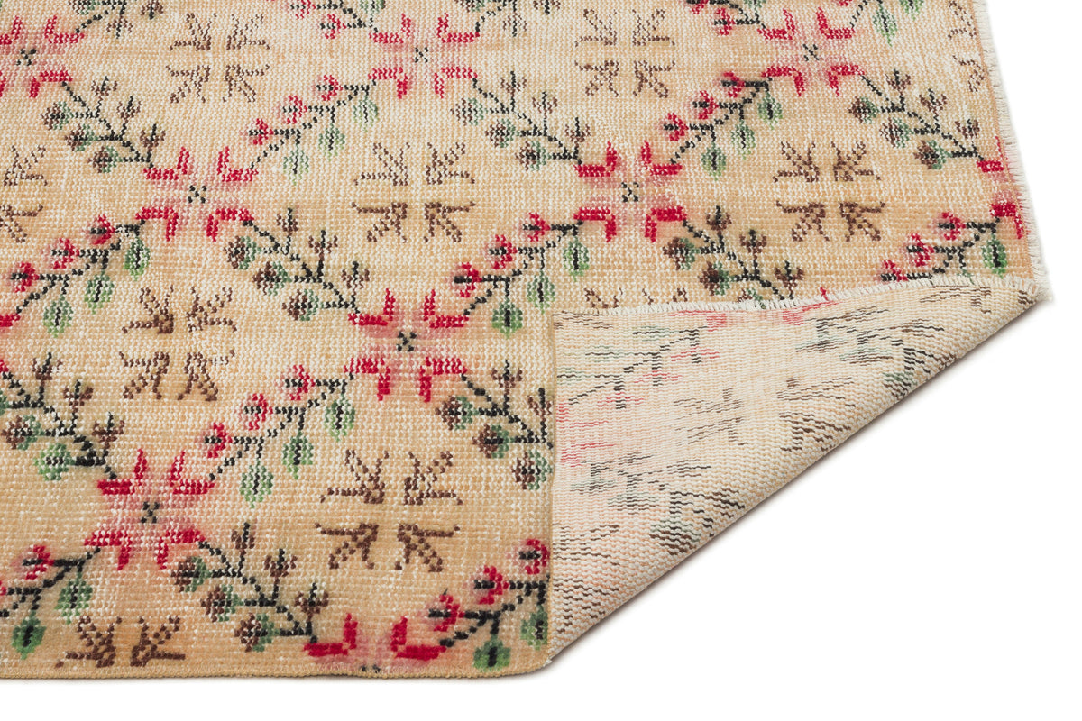 Beige vintage-style rug with a repeating floral pattern in red and green, featuring a folded corner showing the backing.